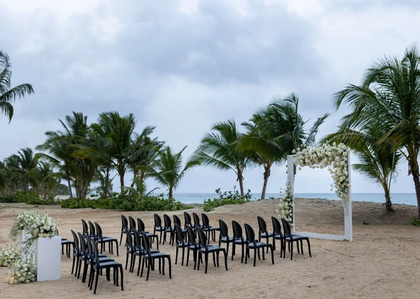 A beach wedding setup with rows of black chairs facing a floral arch, with the ocean in the background.