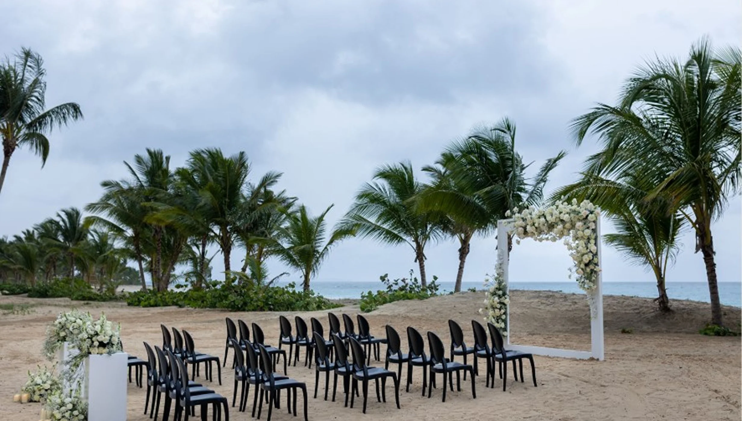 A beach wedding setup with rows of black chairs facing a floral arch, with the ocean in the background.