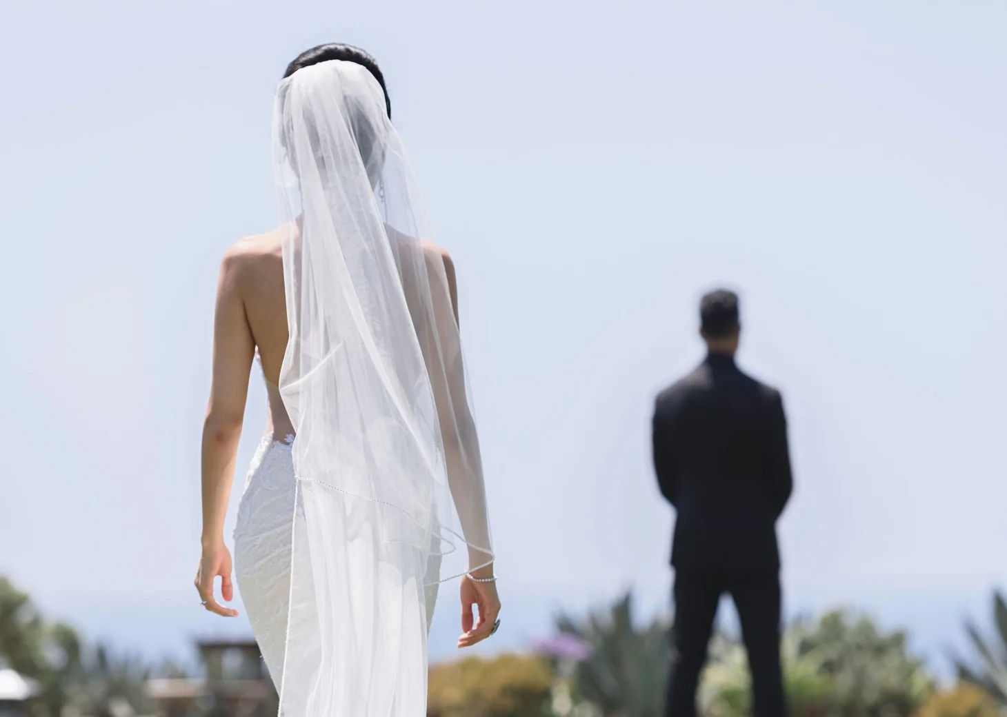 A bride in a flowing white dress and veil walks towards the groom, who is out of focus in the background.