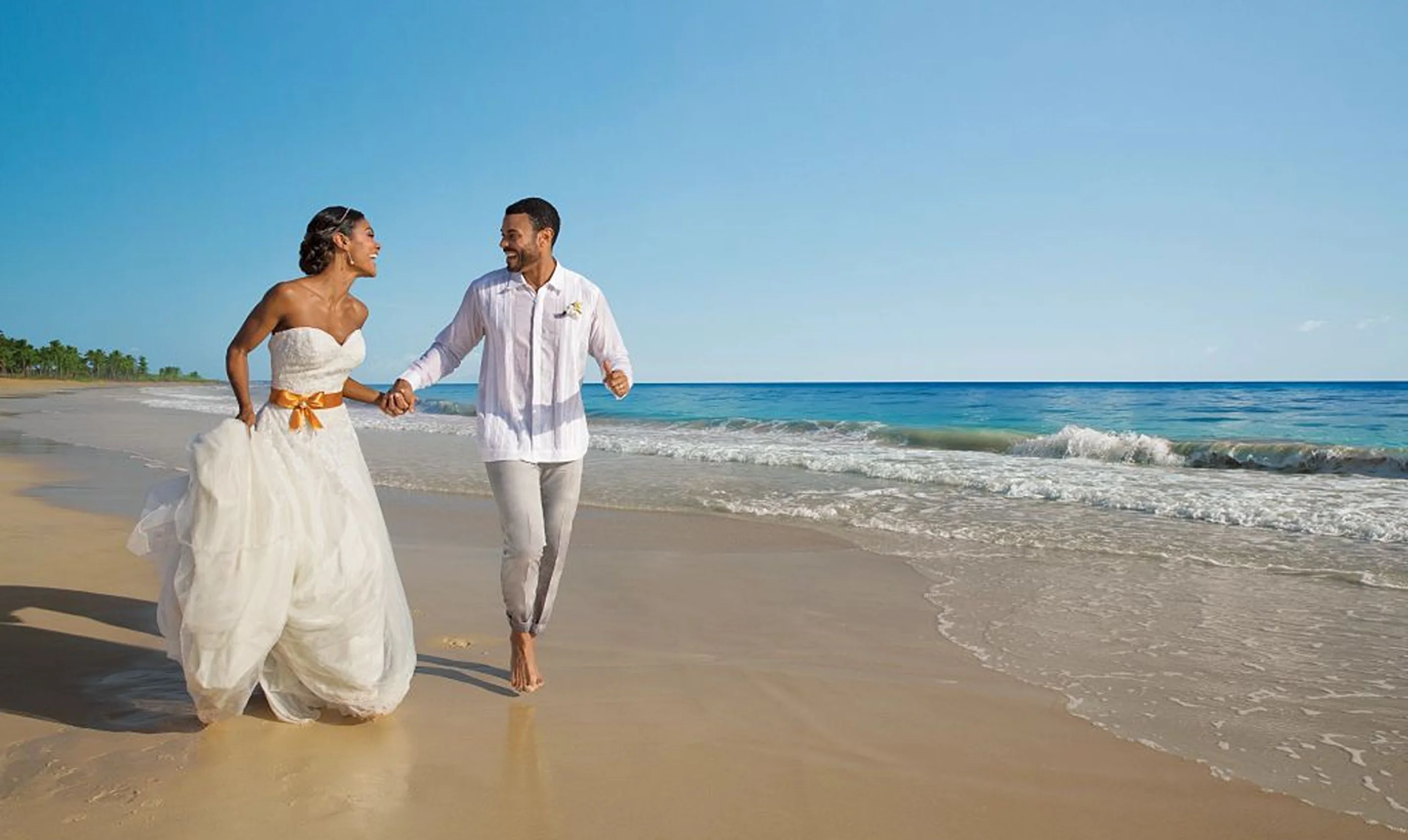 A joyful bride and groom, in wedding attire, holding hands and running along the beautiful sandy beach.