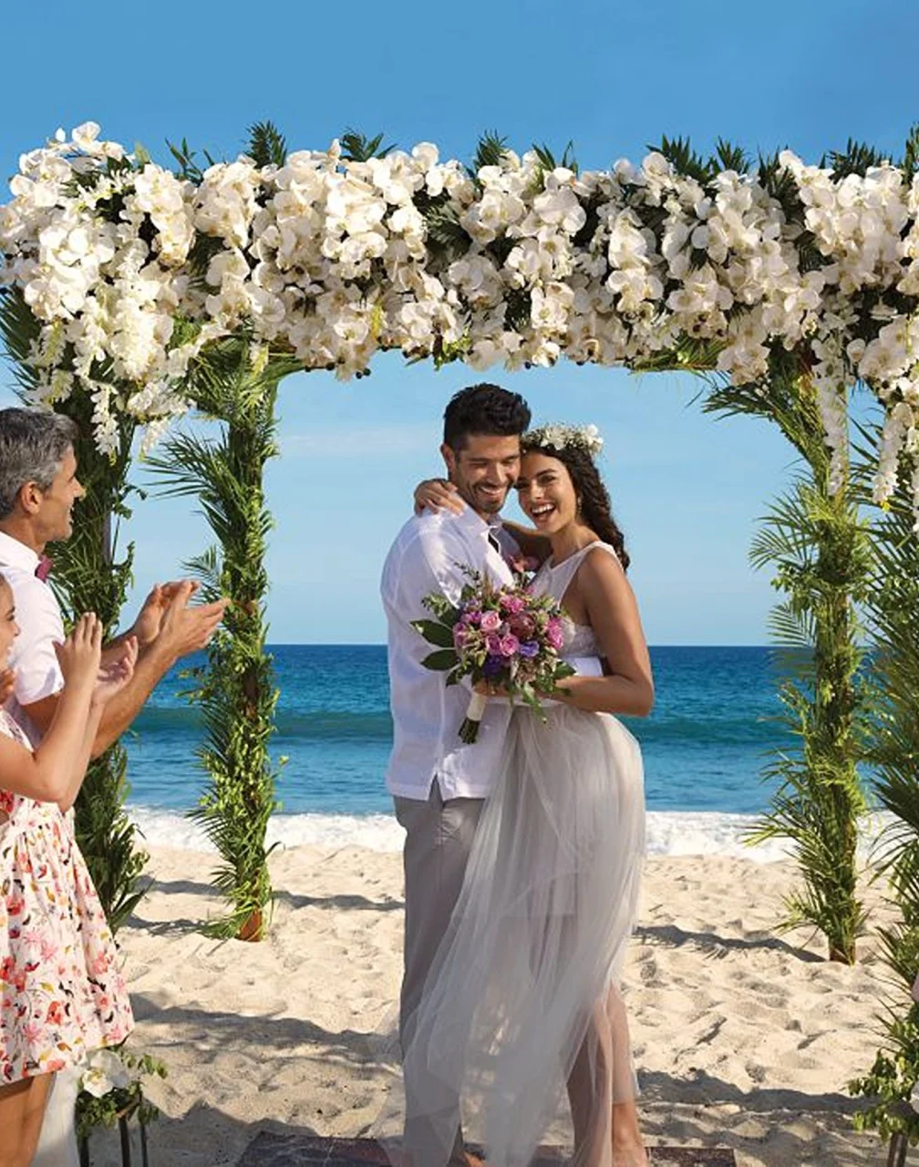 A newly married couple and their happy guests celebrating on a sunlit beach under a beautiful floral arch.