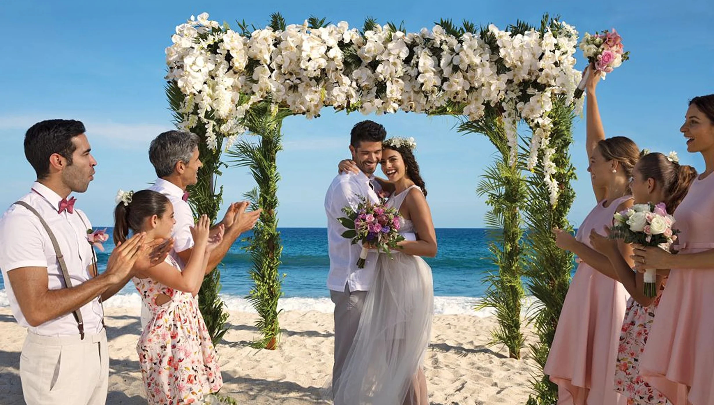 A newly married couple and their happy guests celebrating on a sunlit beach under a beautiful floral arch.