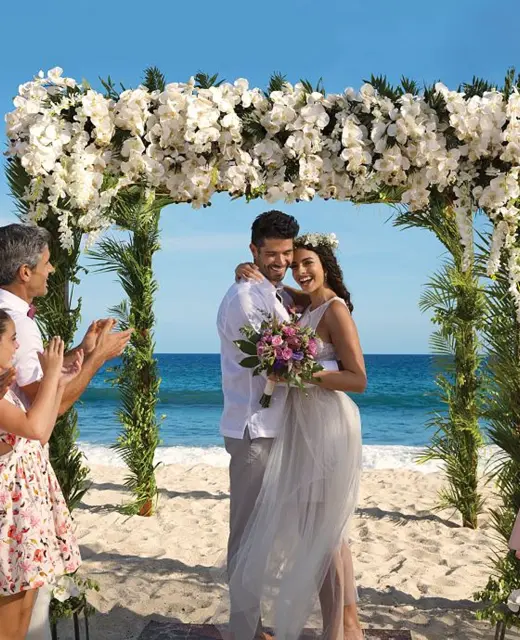 A newly married couple and their happy guests celebrating on a sunlit beach under a beautiful floral arch.
