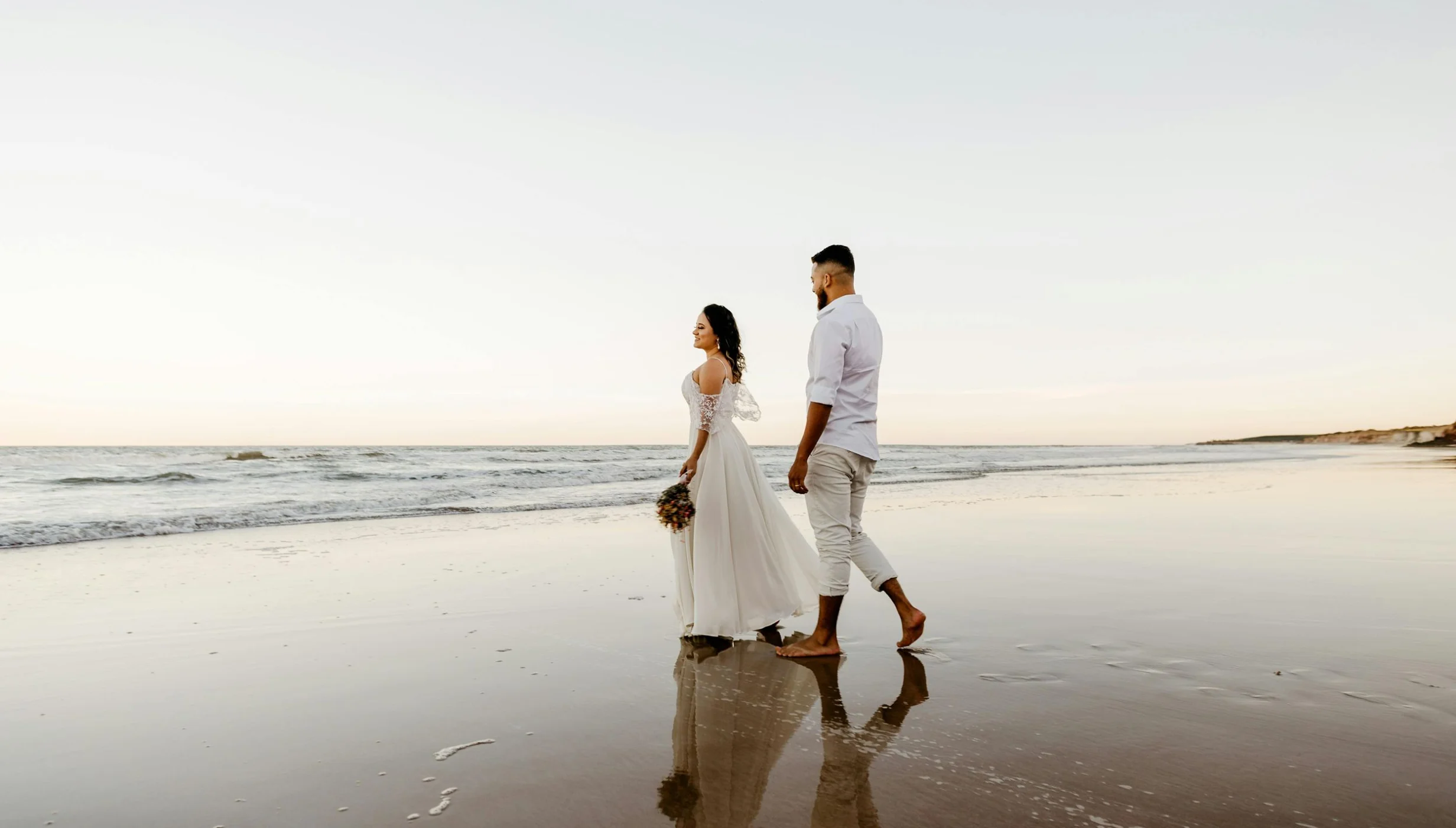 A bride and groom walking barefoot on a wet, sandy beach, their reflections visible in the water.