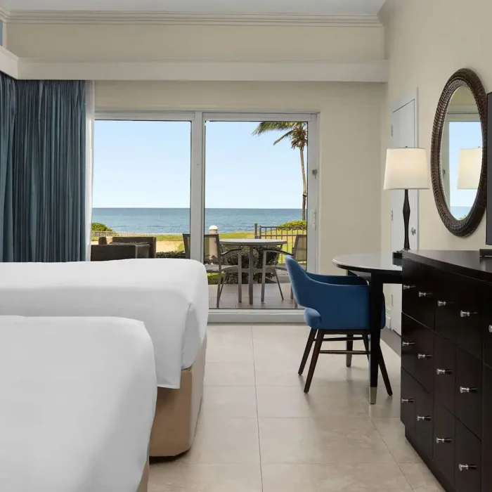 An Oceanfront Walkout Two Queens room with two beds and a walkout patio with a table and chairs by the ocean.