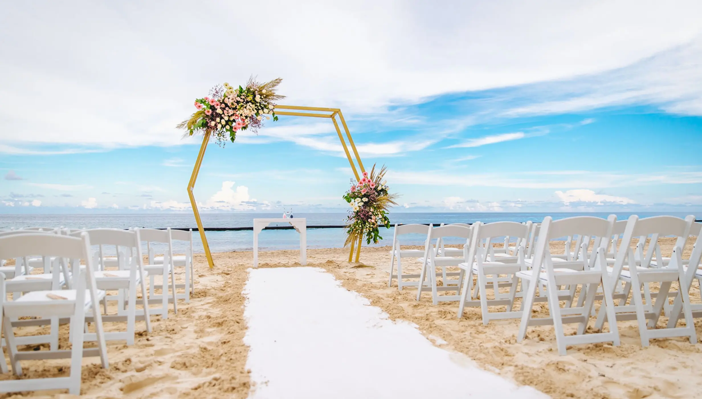 Beachfront wedding ceremony with a gold hexagonal arch and white chairs at Dreams Sapphire, Riviera Maya