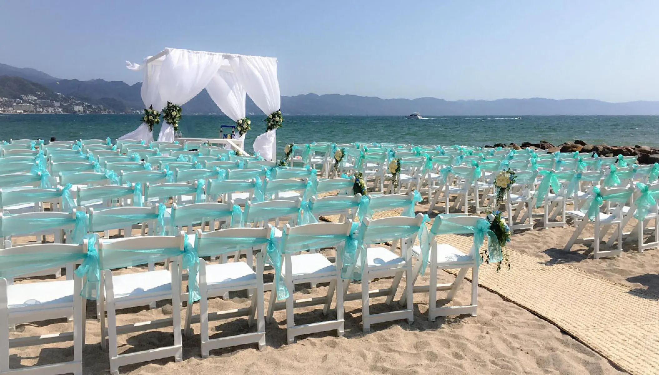 Beach wedding ceremony at Fiesta Americana Puerto Vallarta with white draped arch and chairs with turquoise sashes