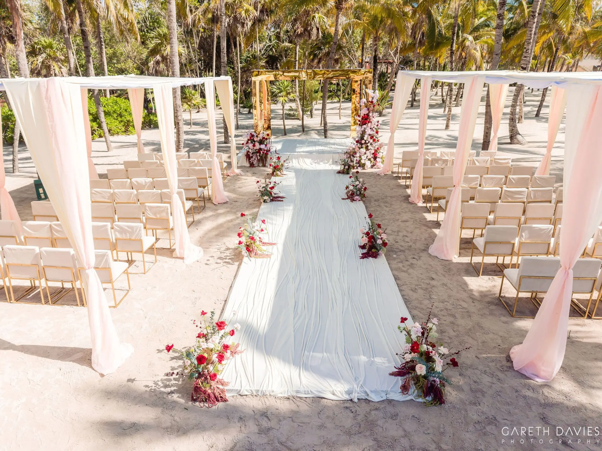 Beachfront wedding ceremony setup with pink draped canopy and red floral aisle at Grand Palladium Riviera Maya