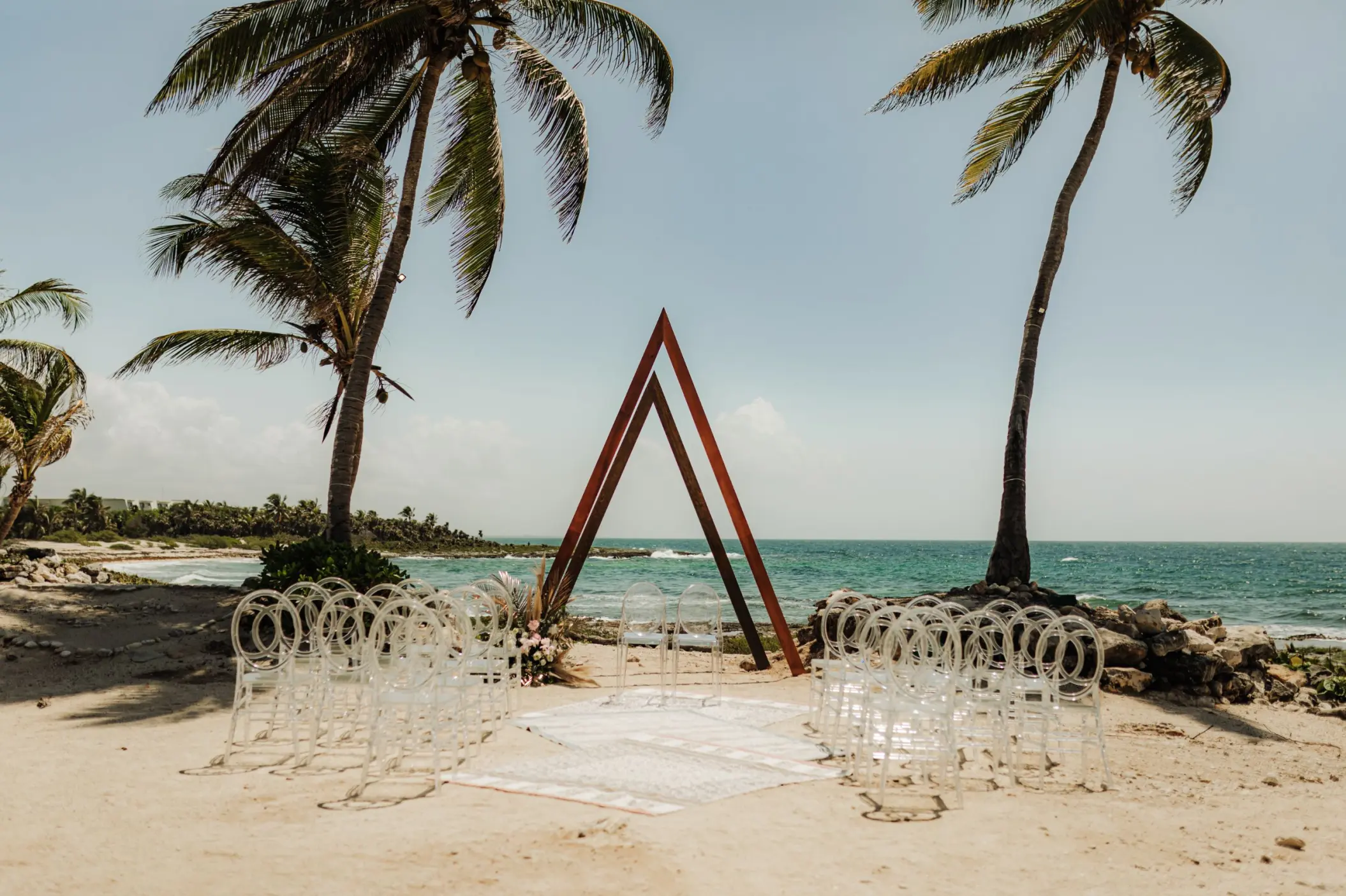 Beachfront wedding ceremony with a triangular arch and clear chairs at Grand Sirenis, Riviera Maya