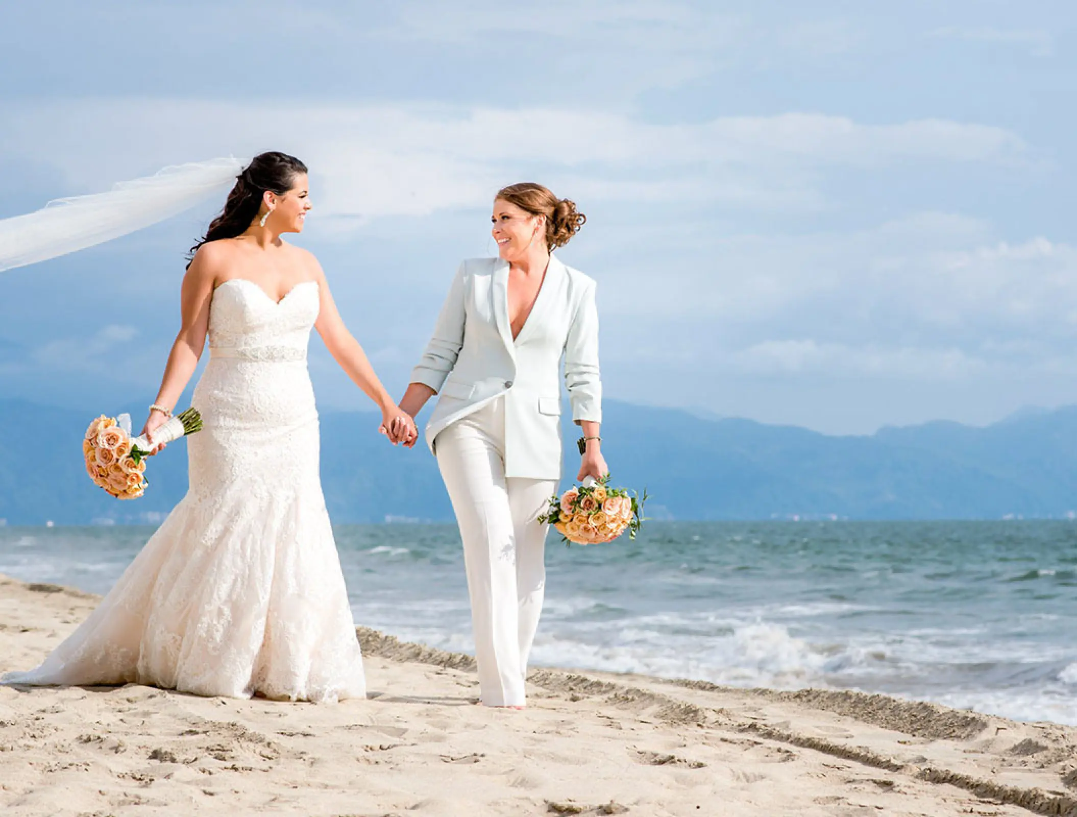 Two brides holding hands and walking on the beach at Grand Velas Riviera Nayarit, Puerto Vallarta