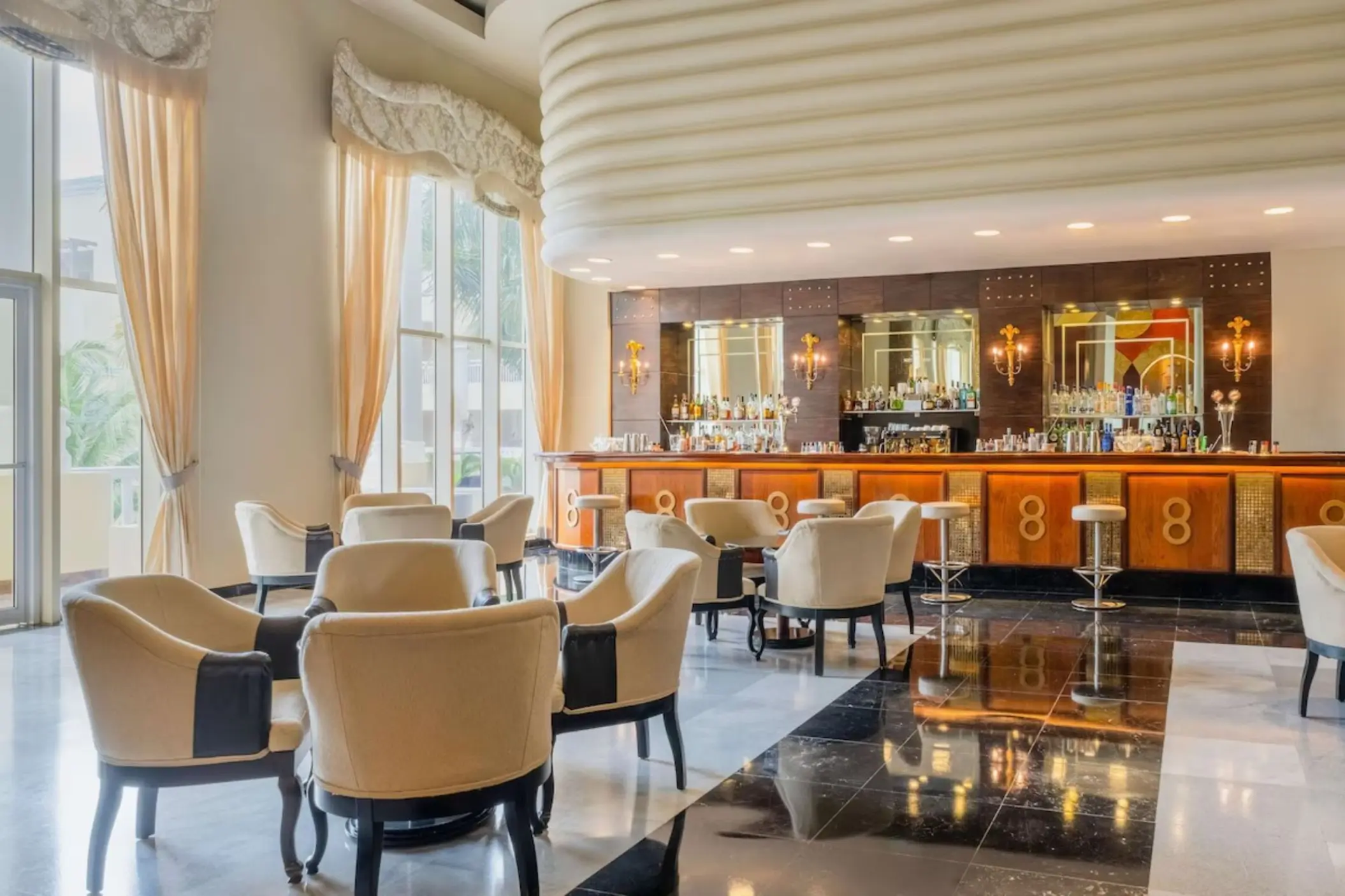 Elegant indoor bar and lounge area with cream chairs and marble floors at JOIA Rose Hall, Montego Bay