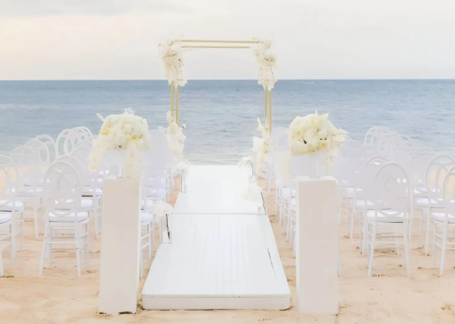 Beachfront wedding ceremony with white chairs and floral arch at JOIA Rose Hall by Iberostar, Montego Bay