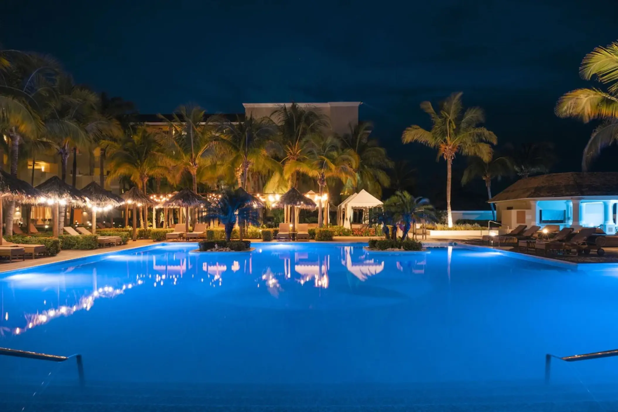 Illuminated swimming pool at night surrounded by palm trees at JOIA Rose Hall, Montego Bay