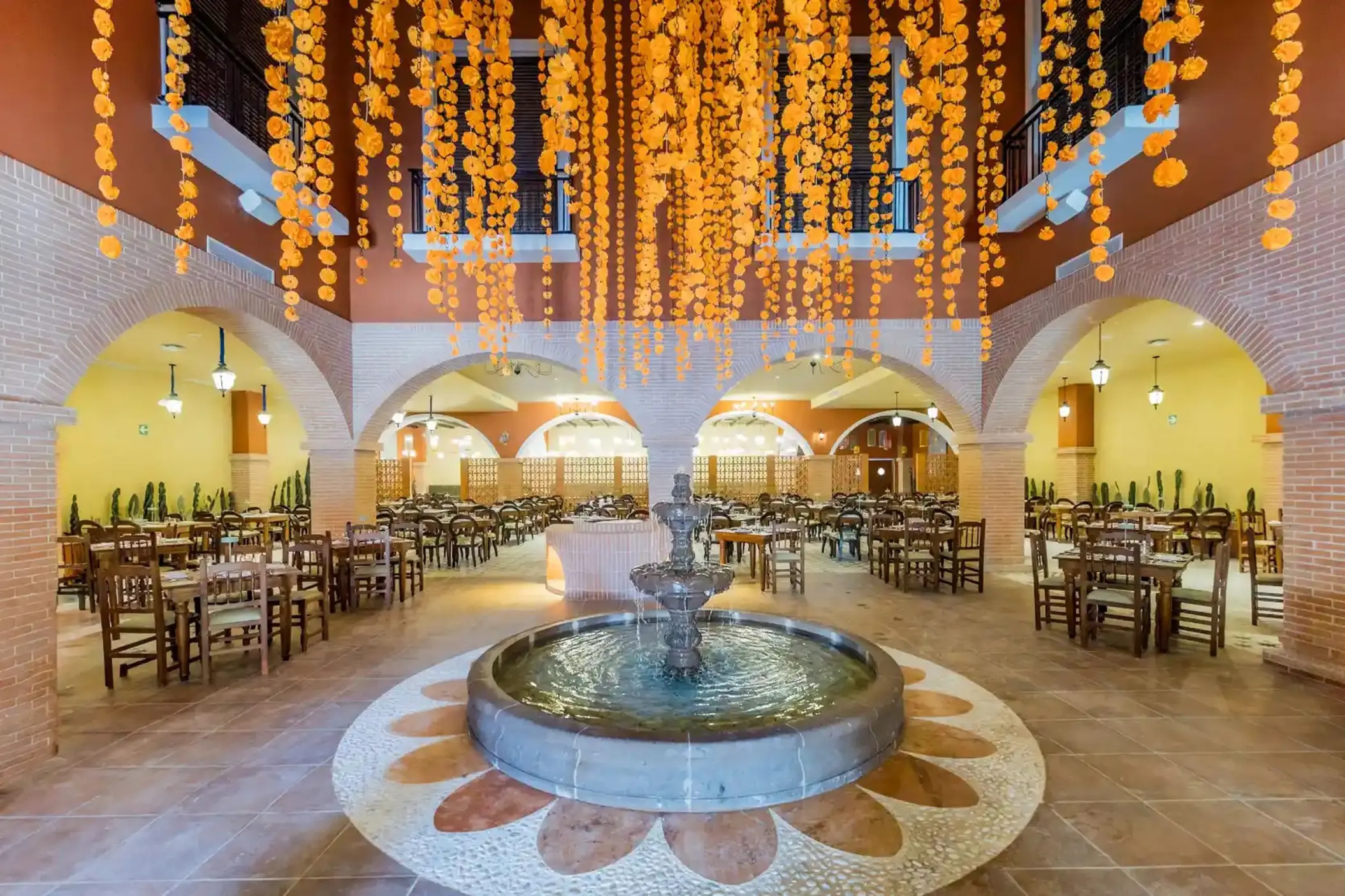 Indoor restaurant with a central stone fountain and hanging orange floral garlands at Majestic Mirage, Cancun