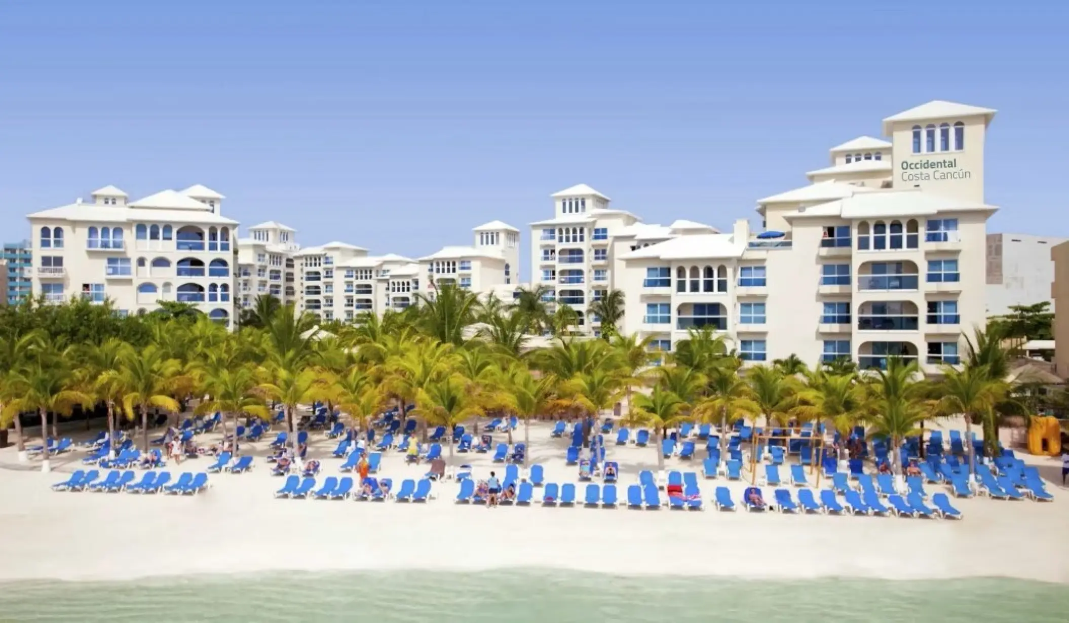 White resort buildings and palm trees fronting a beach with blue lounge chairs at Occidental Costa Cancun