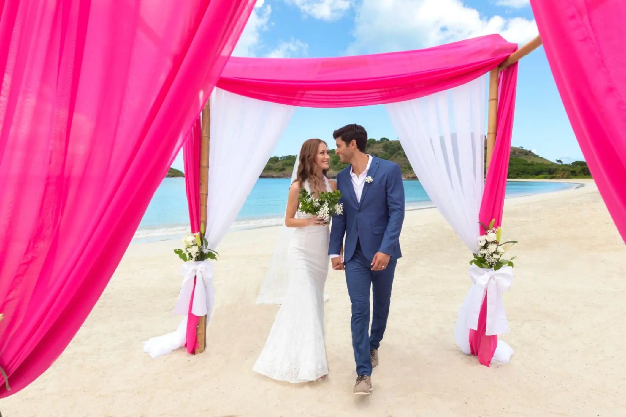 Bride and groom under a pink and white beach ceremony arch at Royalton Antigua, Five Islands Village