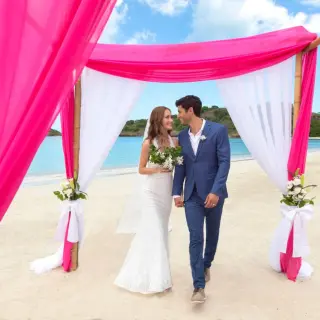 Bride and groom under a pink and white beach ceremony arch at Royalton Antigua, Five Islands Village