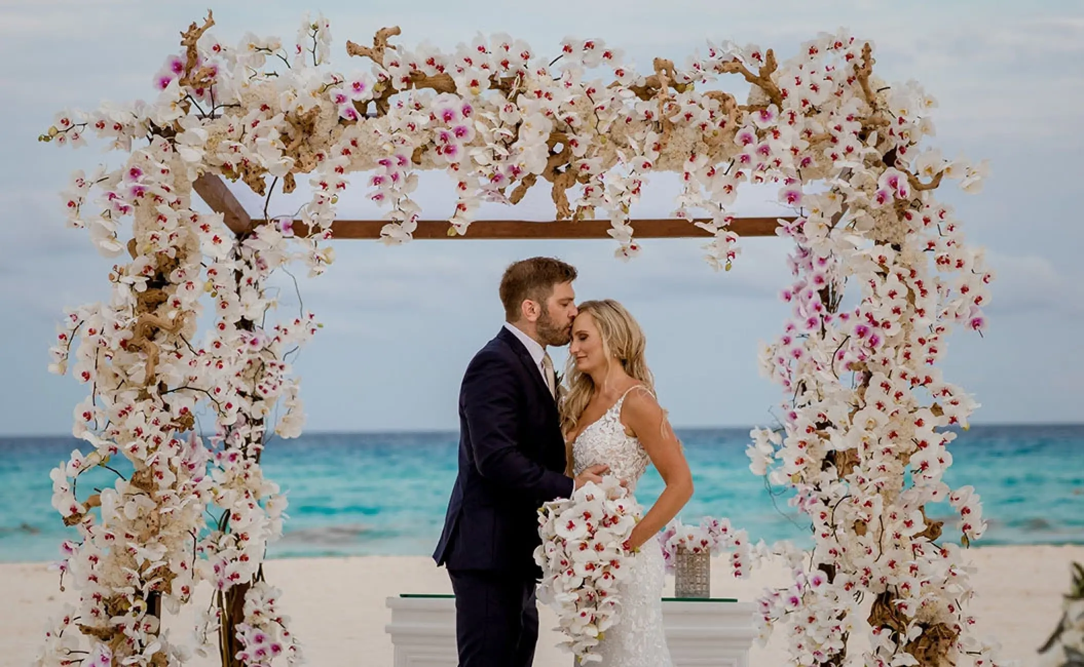 The groom kissing the bride's forehead under a floral arch at Secrets Playa Blanca, Cancun