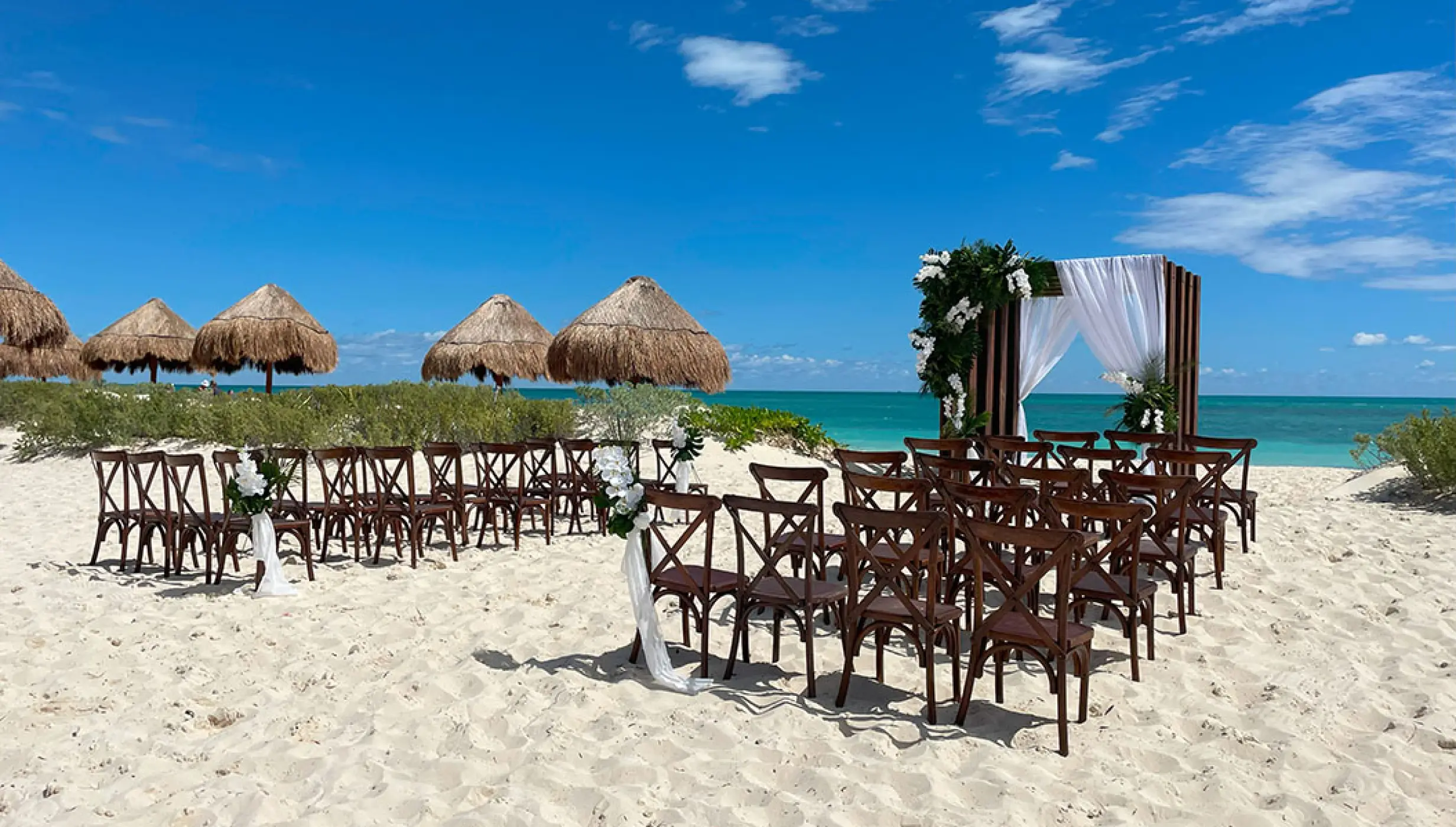 Beachfront wedding ceremony with wooden cross-back chairs and a floral arch at Secrets Playa Blanca, Cancun