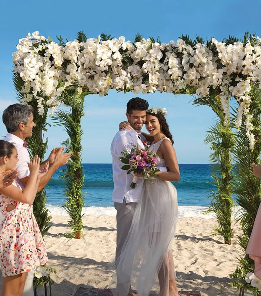 A newly married couple and their happy guests celebrating on a sunlit beach under a beautiful floral arch.
