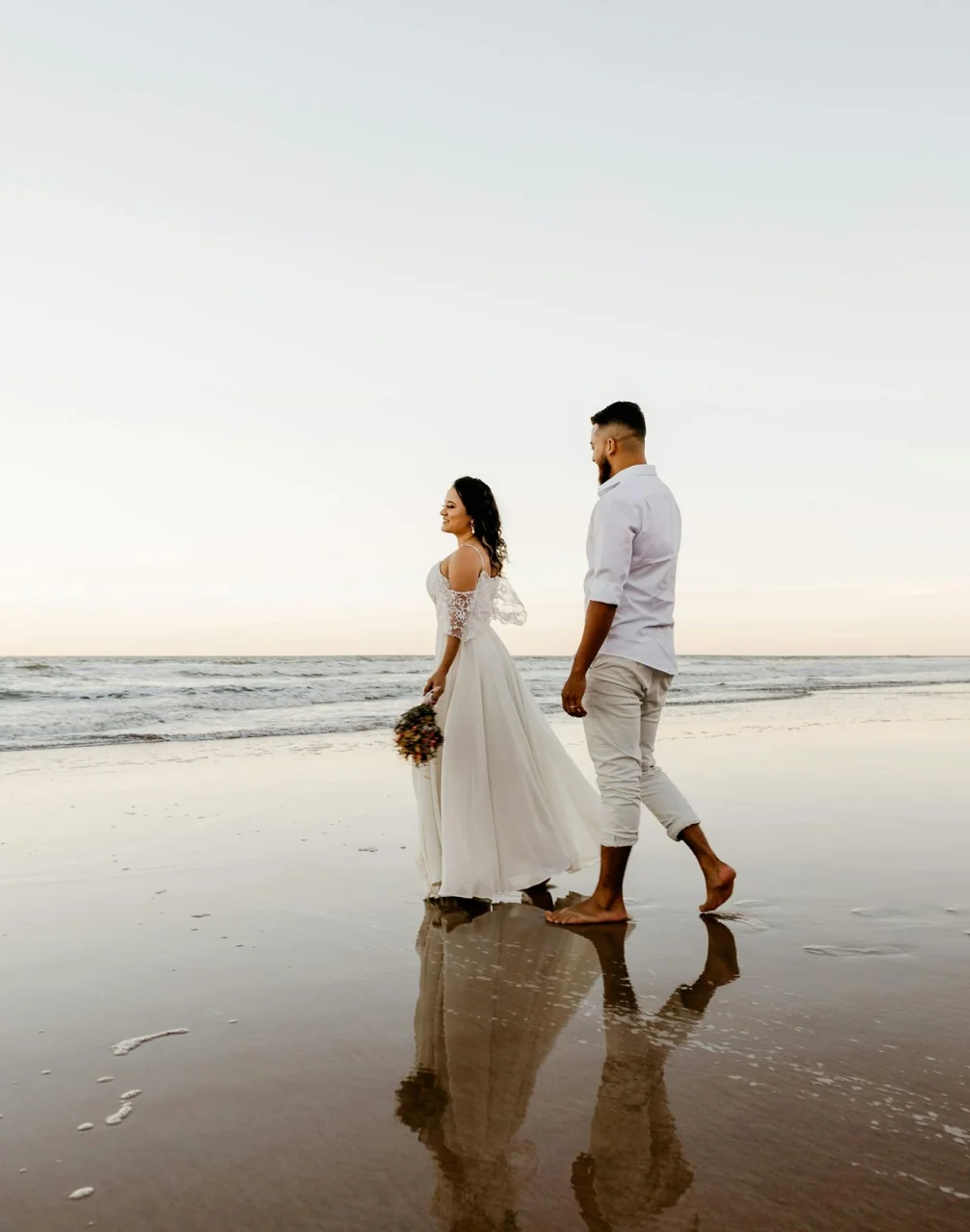 A bride and groom walking barefoot on a wet, sandy beach, their reflections visible in the water.