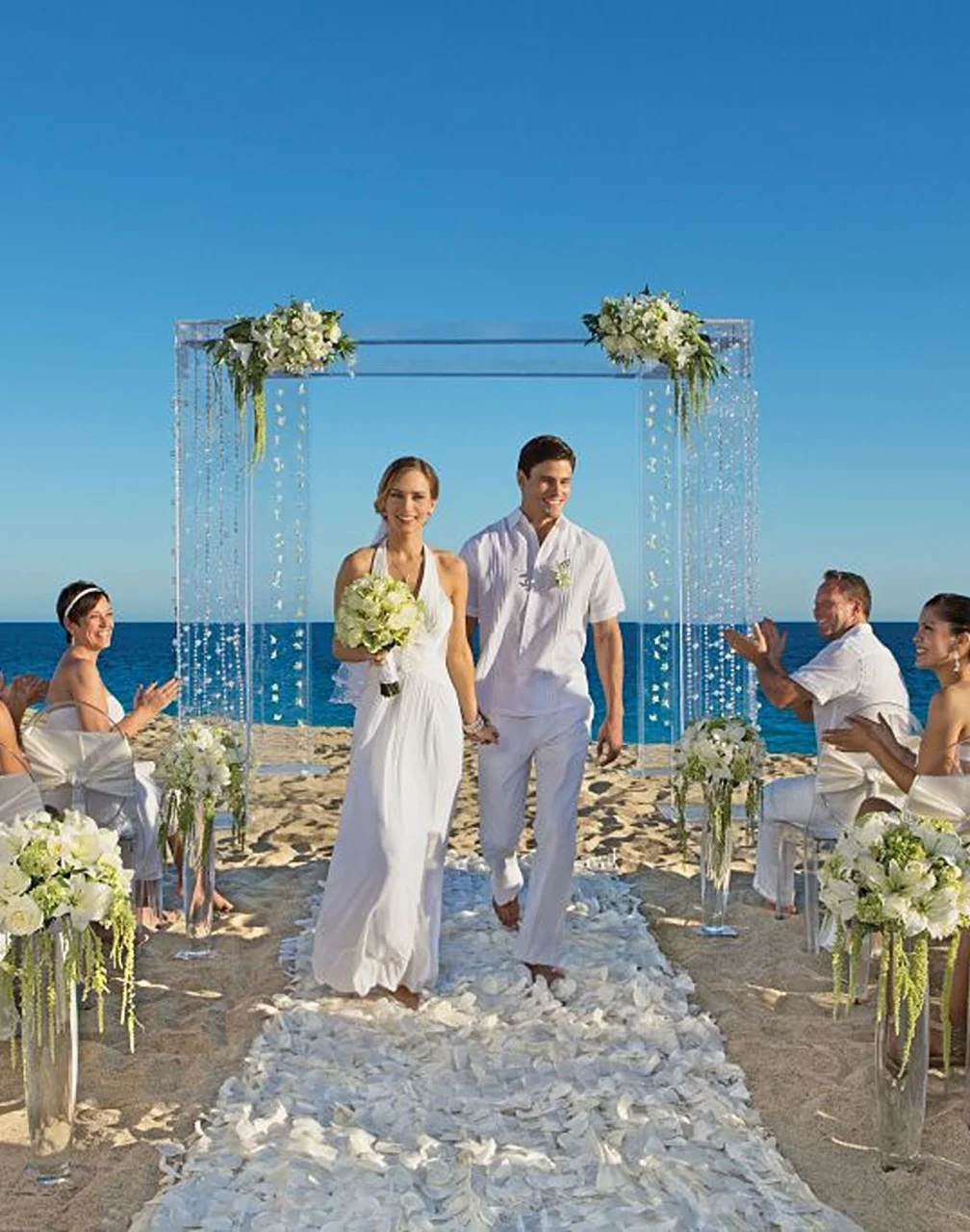 A happy couple walking down a petal-strewn aisle on the beach, surrounded by applauding guests.