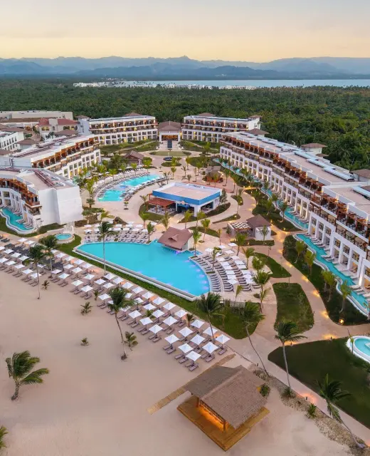 An aerial view of the Secrets Playa Esmeralda resort at sunset, with pools, beach, and lush trees.