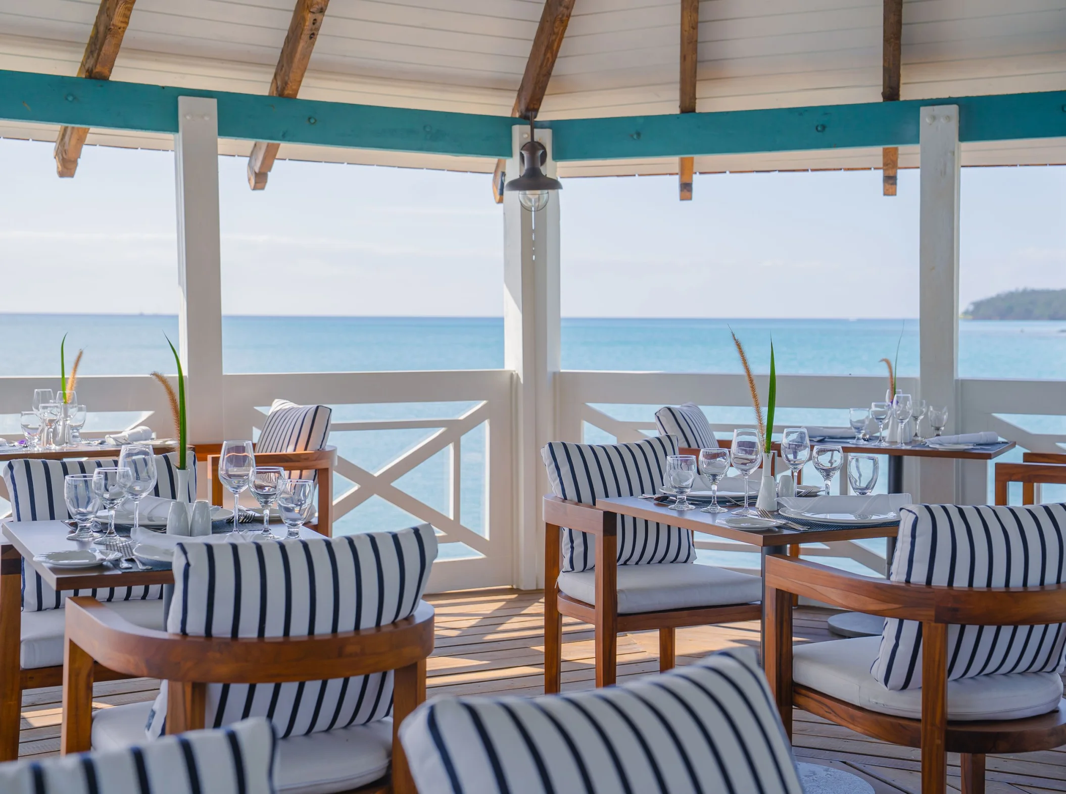 An open-air restaurant with black and white striped chairs and tables set overlooking the clear blue ocean.