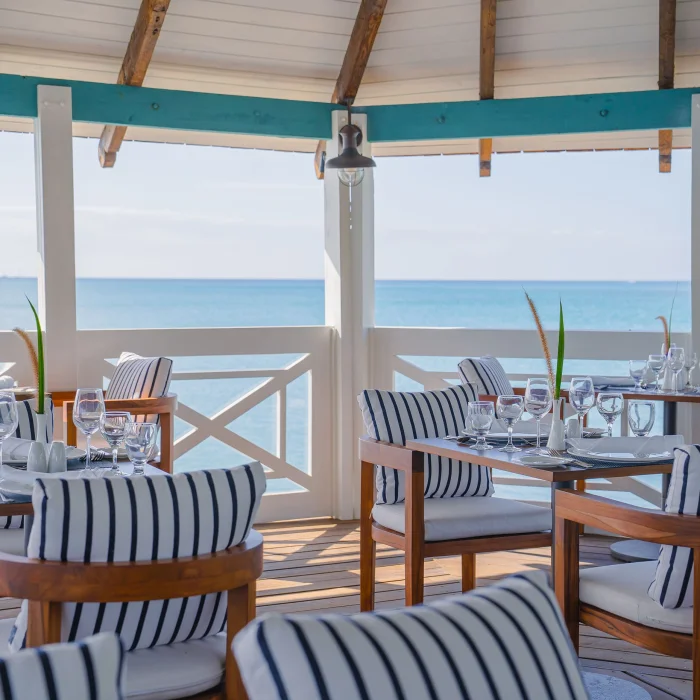 An open-air restaurant with black and white striped chairs and tables set overlooking the clear blue ocean.