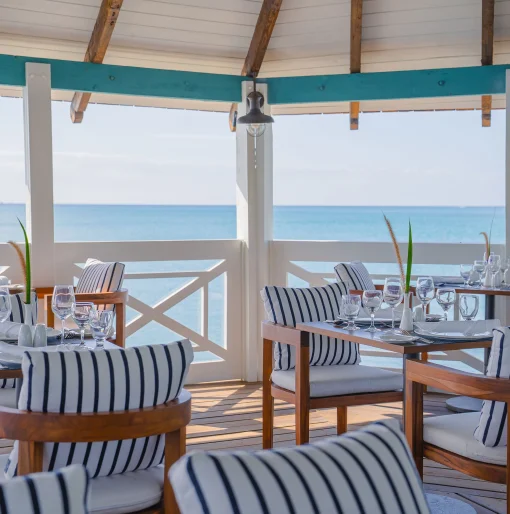 An open-air restaurant with black and white striped chairs and tables set overlooking the clear blue ocean.