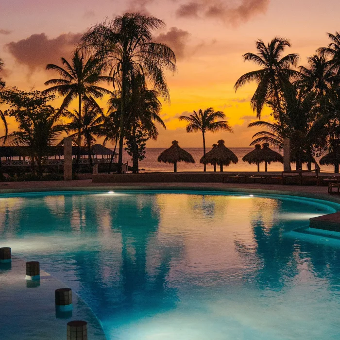 A stunning resort pool at sunset, surrounded by palm trees and tiki huts, with an orange sky.
