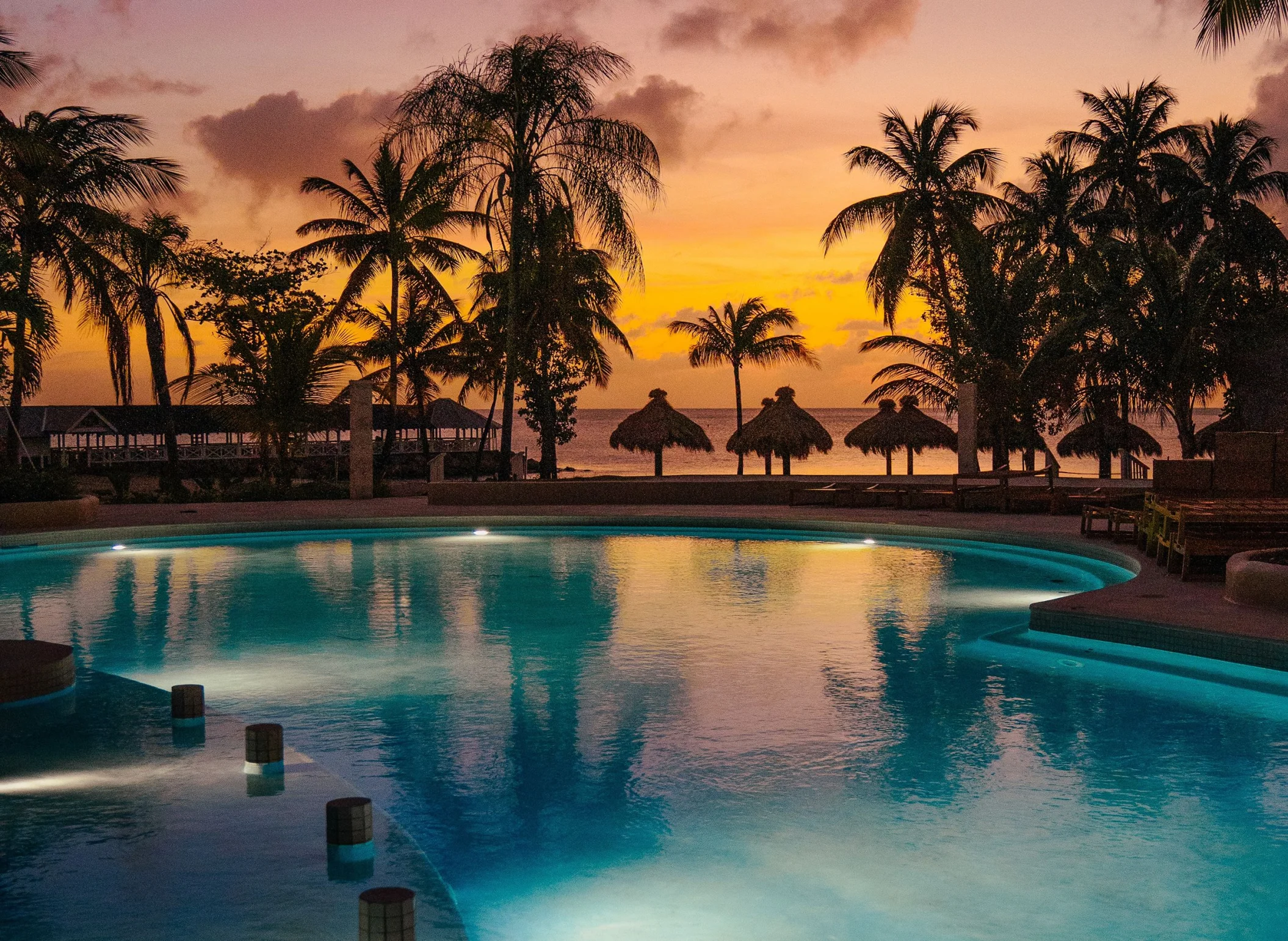A stunning resort pool at sunset, surrounded by palm trees and tiki huts, with an orange sky.