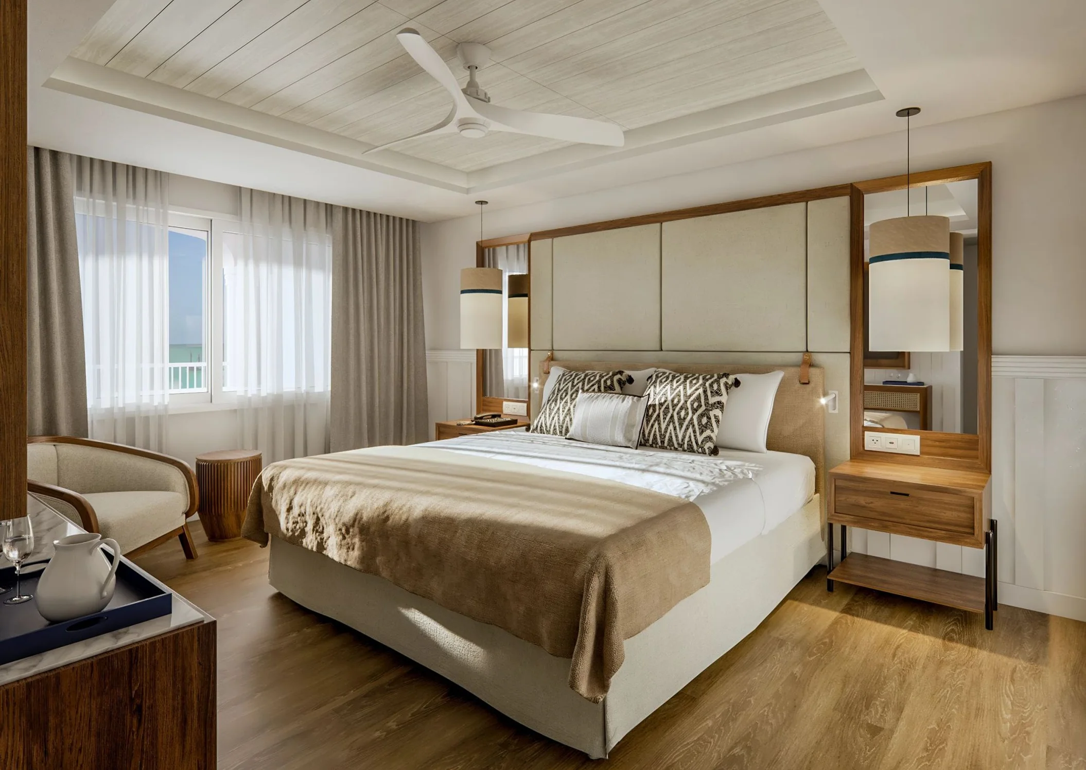 A luxurious hotel bedroom with a large bed, ceiling fan, and bright windows looking out to the sea.