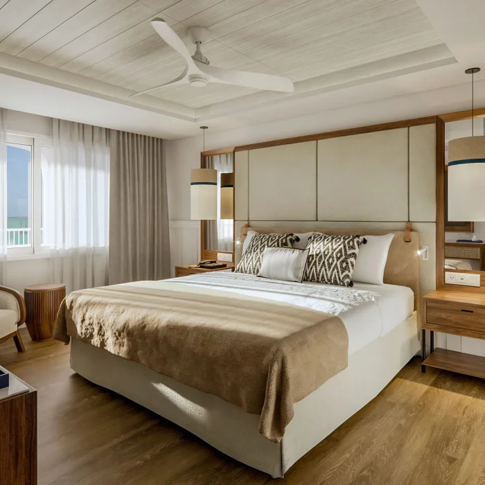 A luxurious hotel bedroom with a large bed, ceiling fan, and bright windows looking out to the sea.