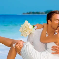 A joyful wedding couple on a beach, with the groom carrying the bride against a blue ocean backdrop.