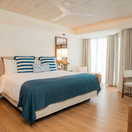 A bright hotel room with a large king bed, ceiling fan, and sheer curtains leading to a view.