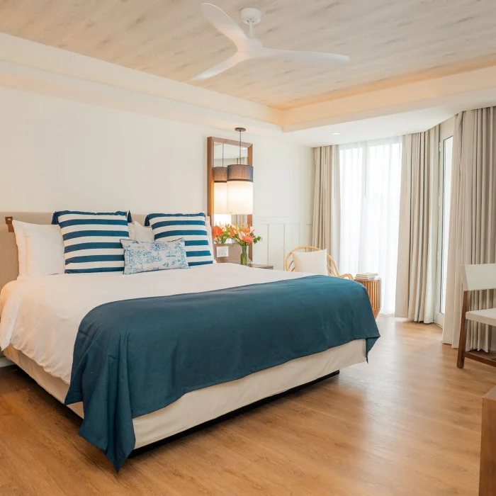 A bright hotel room with a large king bed, ceiling fan, and sheer curtains leading to a view.