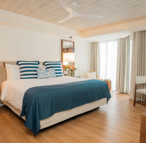 A bright hotel room with a large king bed, ceiling fan, and sheer curtains leading to a view.