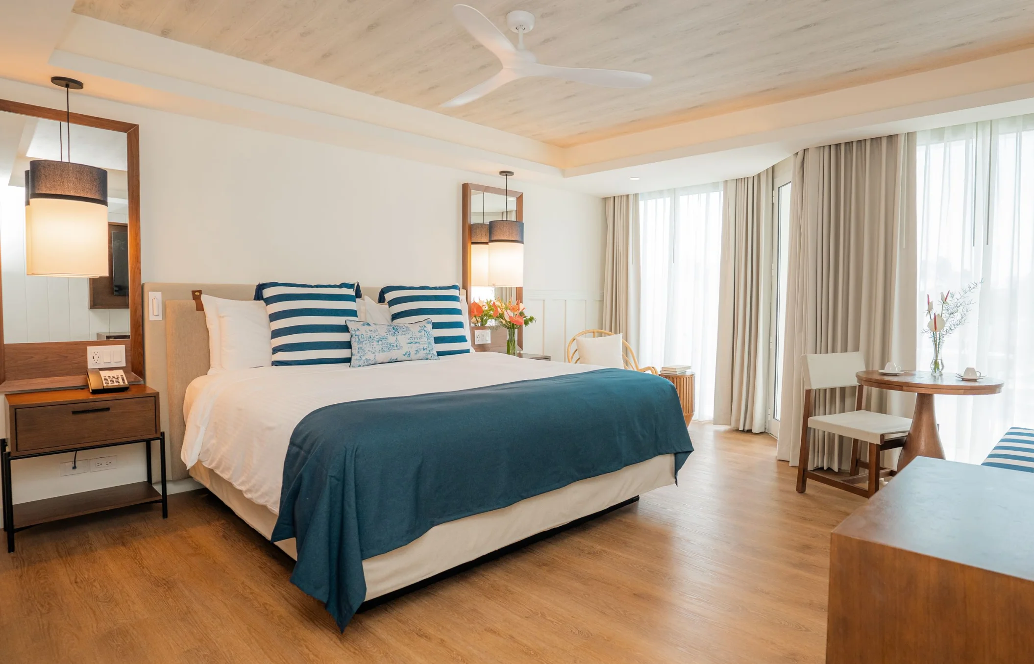 A bright hotel room with a large king bed, ceiling fan, and sheer curtains leading to a view.