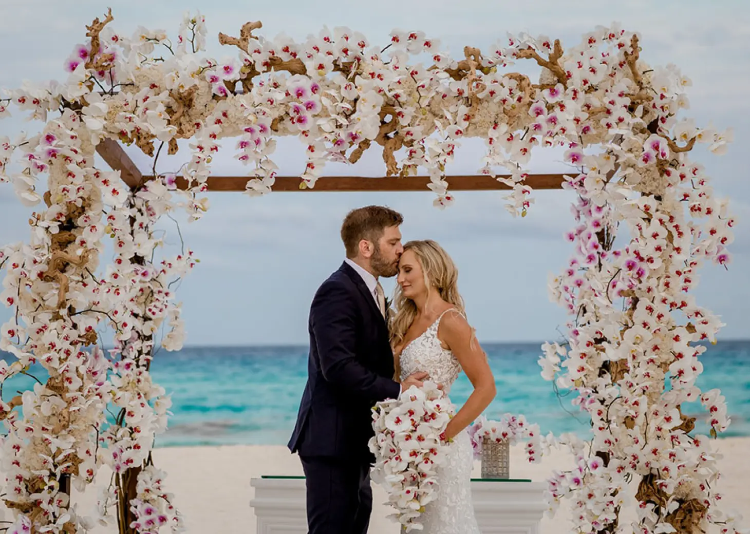The couple sharing a forehead kiss under a floral arch at Secrets The Vine, Cancun