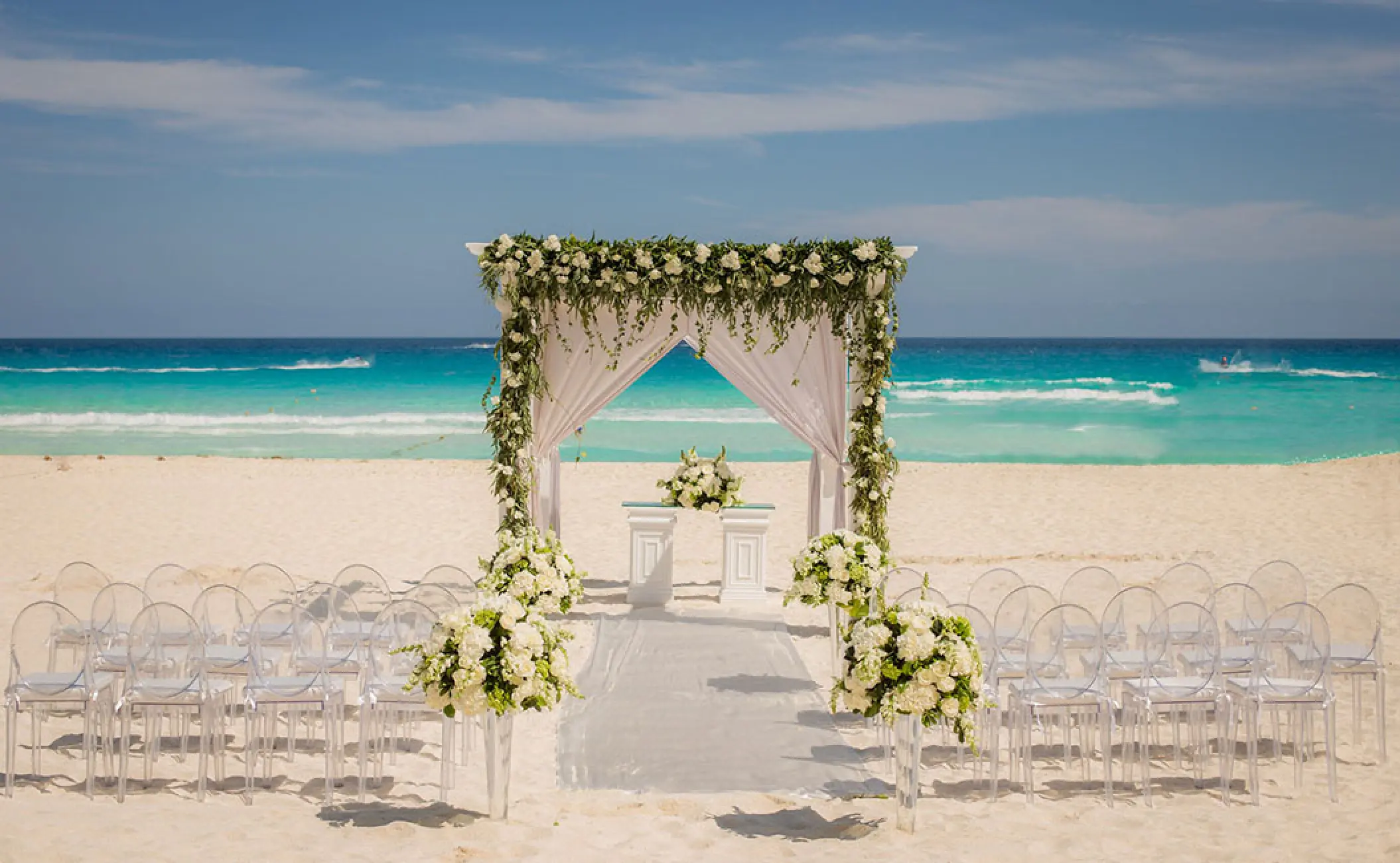 Beachfront wedding ceremony with a floral gazebo and clear chairs at Secrets The Vine, Cancun