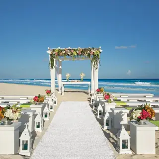 Beachfront wedding ceremony setup with floral gazebo and white benches at Secrets The Vine, Cancun