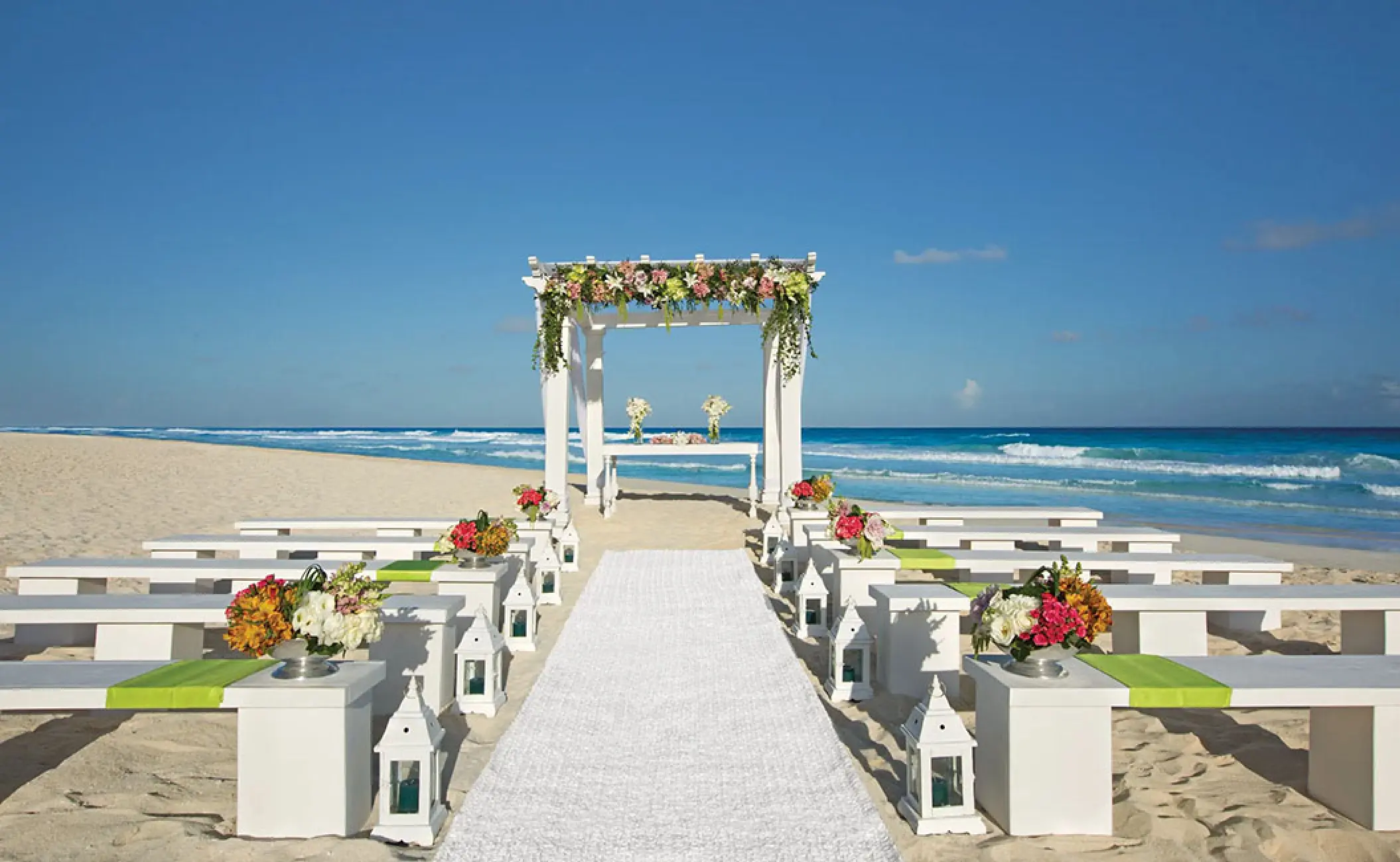 Beachfront wedding ceremony setup with floral gazebo and white benches at Secrets The Vine, Cancun