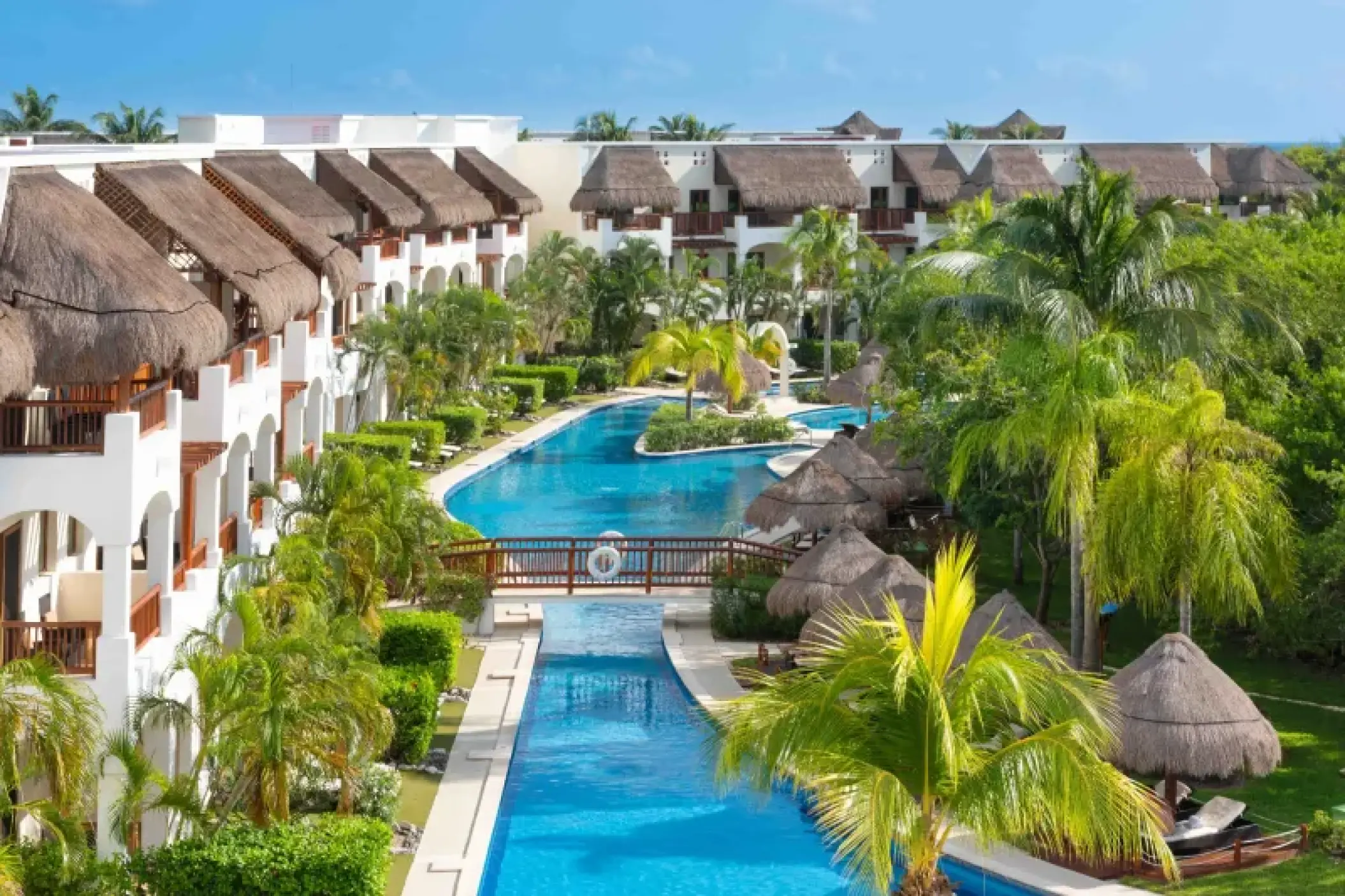 Large winding swimming pool surrounded by thatched-roof buildings and palm trees at Valentin Imperial, Riviera Maya