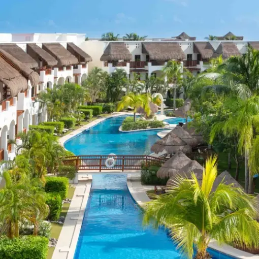 Large winding swimming pool surrounded by thatched-roof buildings and palm trees at Valentin Imperial, Riviera Maya