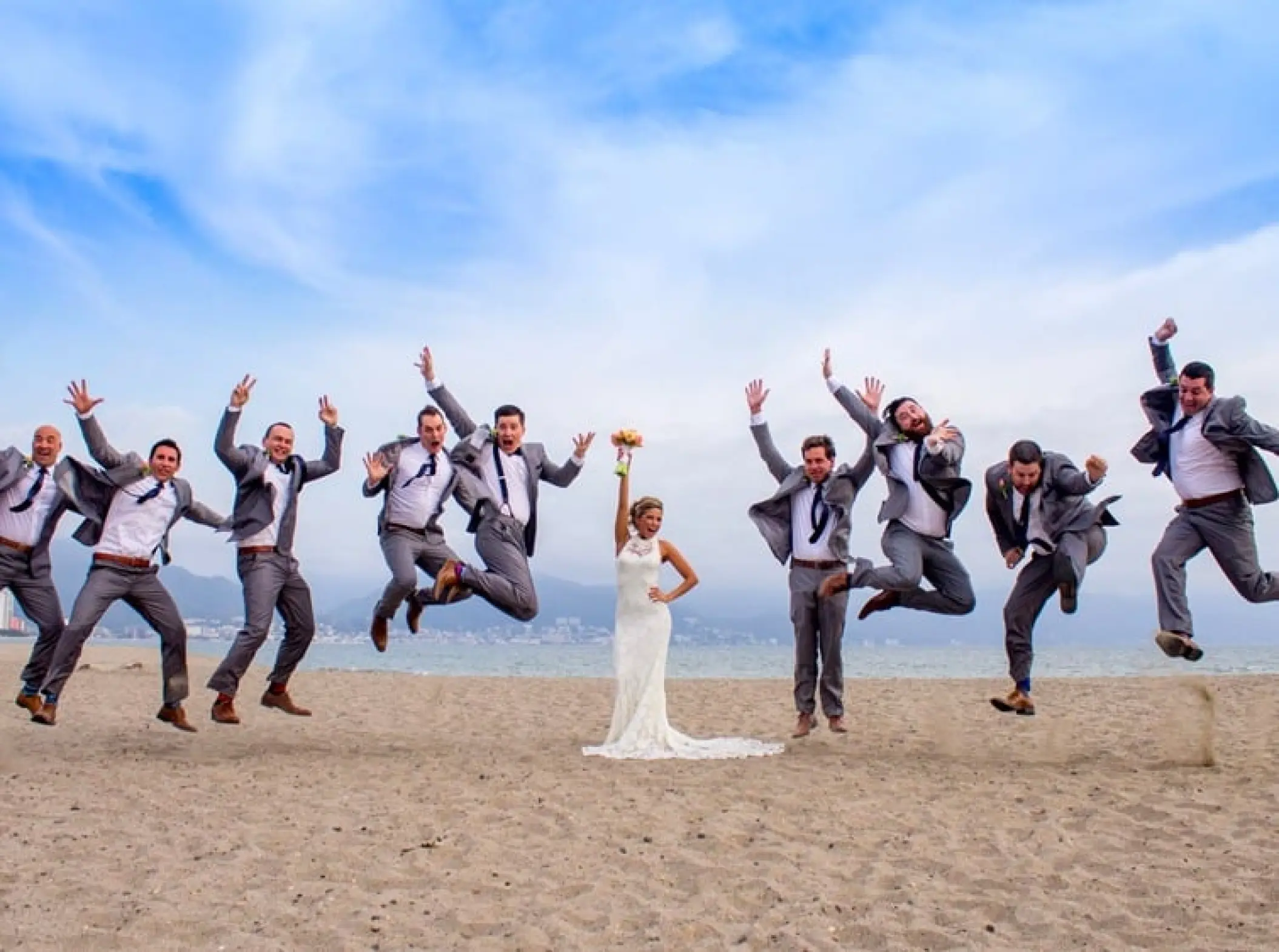Bride standing on the beach with groomsmen jumping in the air at Velas Vallarta, Puerto Vallarta