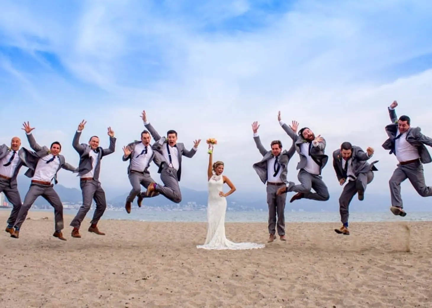 Bride standing on the beach with groomsmen jumping in the air at Velas Vallarta, Puerto Vallarta