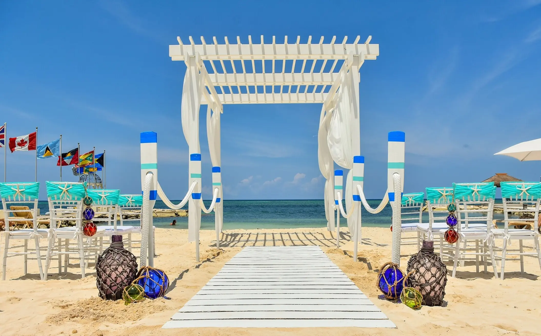 Beachfront wedding ceremony setup with a white pergola and teal chairs at Sandals Montego Bay