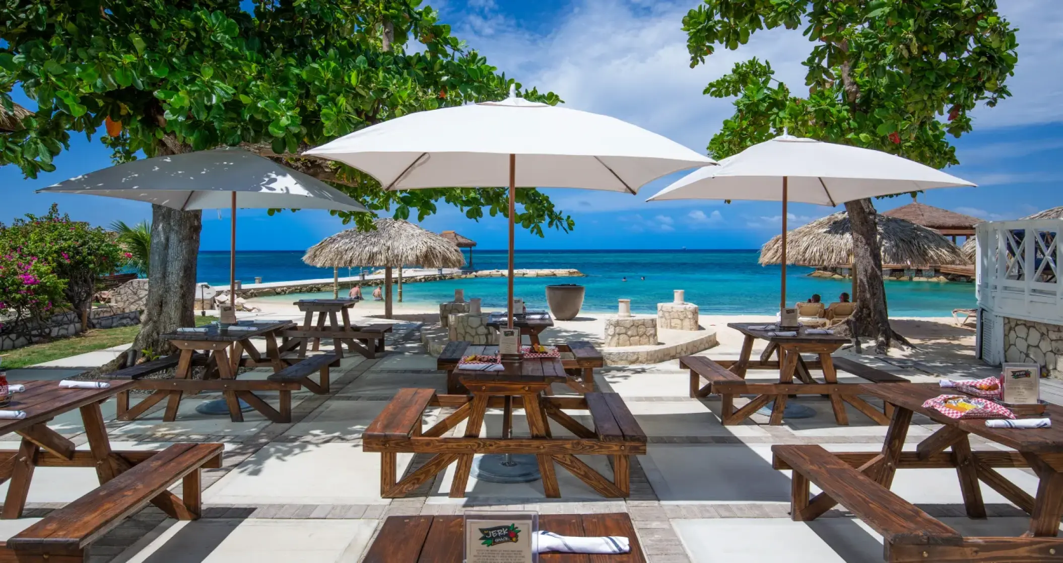 Outdoor beachfront restaurant with picnic tables and white umbrellas at Sandals Montego Bay