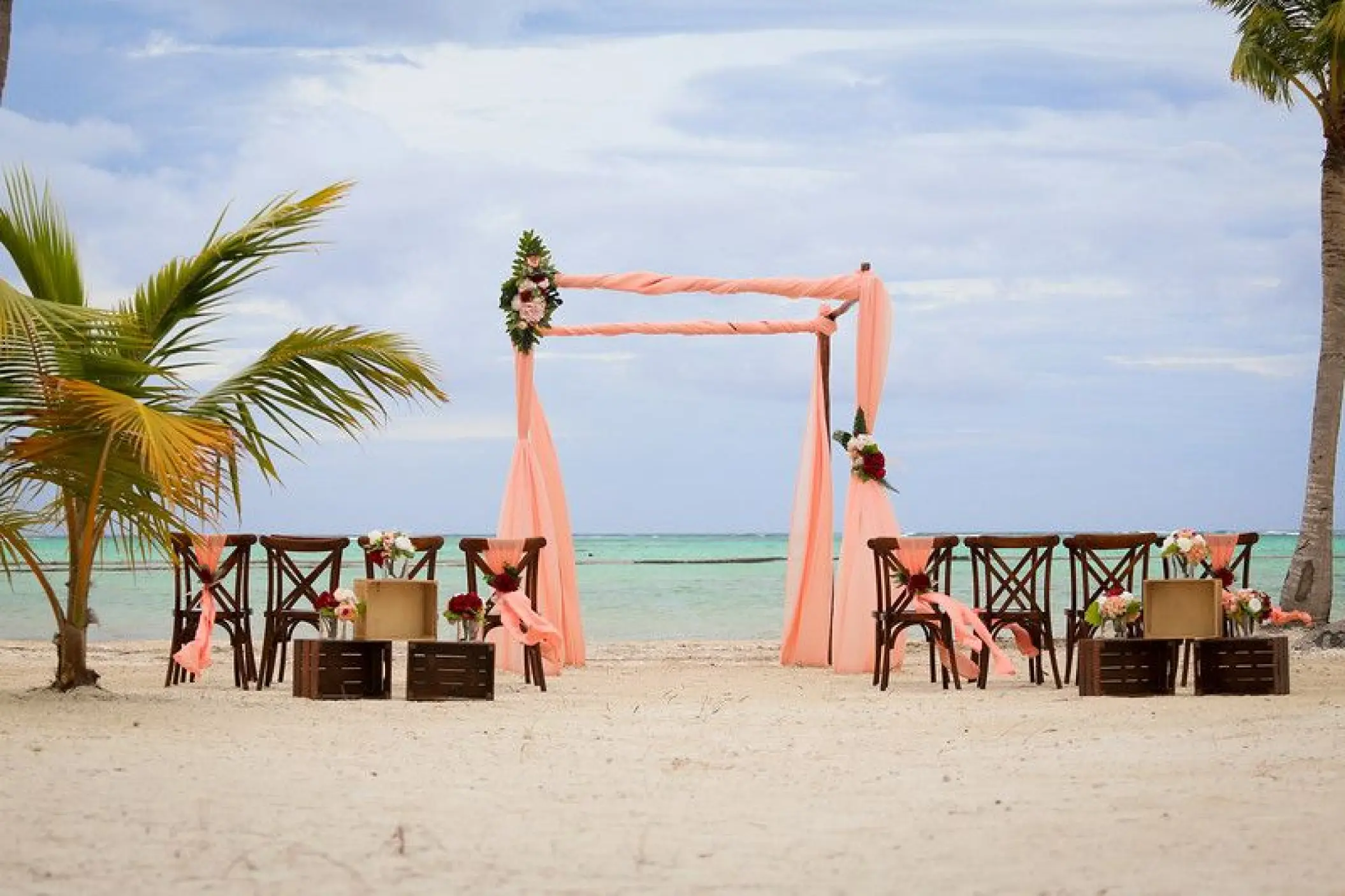 Beachfront wedding ceremony with peach fabric arch and wooden chairs at Secrets Cap Cana, Punta Cana